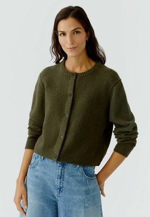 Cardigan vert olive en matériau tricoté doux, avec un col rond, une fermeture à boutons et des poignets côtelés. Associé à un jean en denim bleu clair.