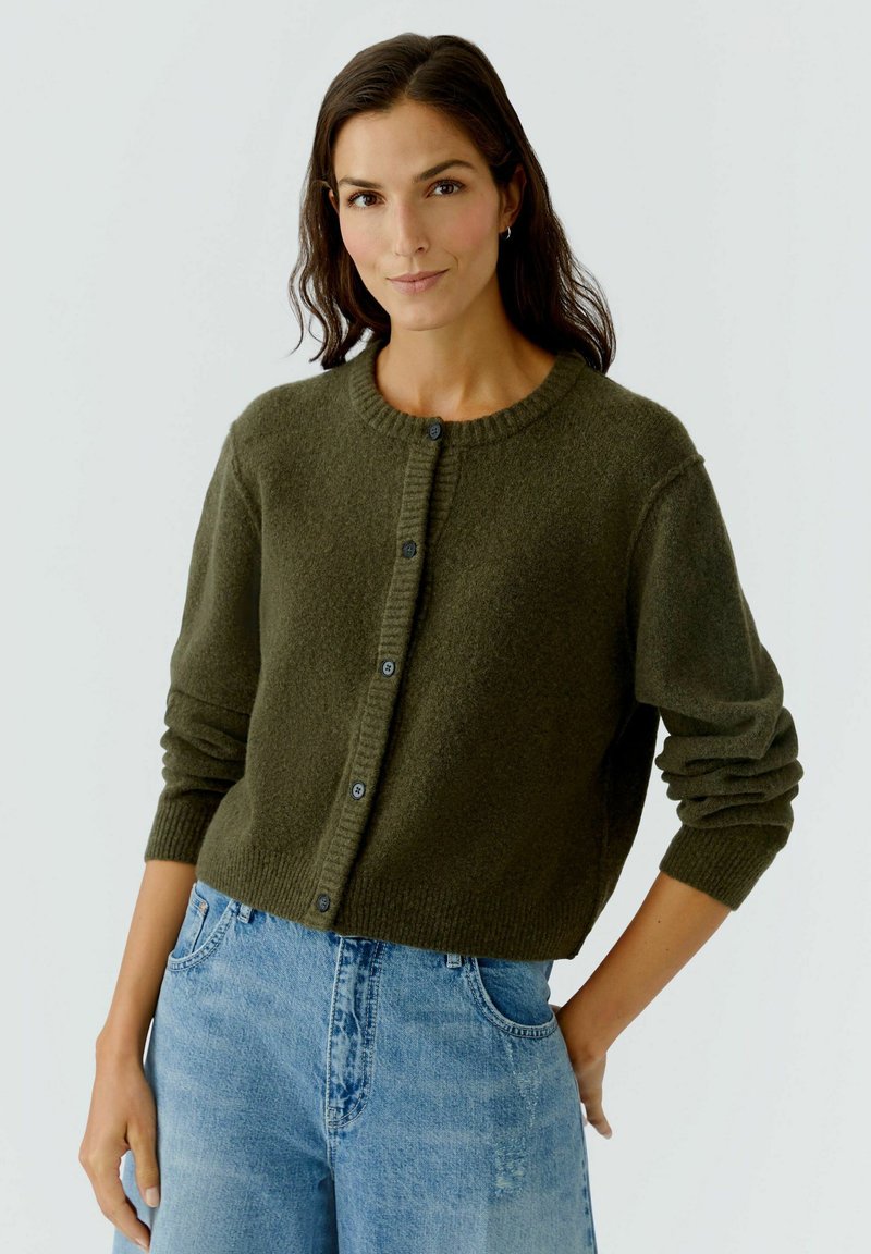 Olivgrüner Cardigan aus weichem Strickmaterial, mit rundem Halsausschnitt, Knopfverschluss und gerippten Bündchen. Kombiniert mit hellblauen Jeans.