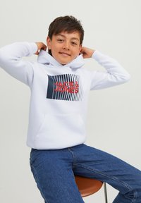 Felpa bianca con tasca frontale, caratterizzata da una grafica "JACK & JONES" in blu e rosso. Abbinata a jeans blu, seduta su uno sgabello.