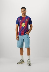Tricou FC Barcelona cu dungi albastre și roșii, prezentând un logo galben Spotify, asortat cu pantaloni scurți din denim deschis și adidași gri.