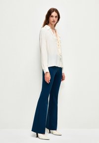 Blusa crema con cuello puntiagudo y lazos en el frente, combinada con pantalones acampanados azules. La modelo lleva zapatos de tacón blanco. Tejido texturizado.