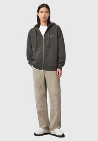 Dunkelgrüner Zip-Hoodie mit verstellbarem Kordelzug, Fronttaschen und weißem Textlogo; kombiniert mit beigen weiten Hosen und weißen Sneakers.