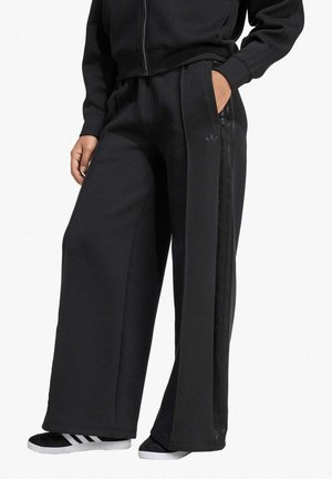 Pantaloni della tuta neri in tessuto morbido, con design a gamba larga, tasche laterali e tre strisce verticali lungo la parte esterna della gamba.