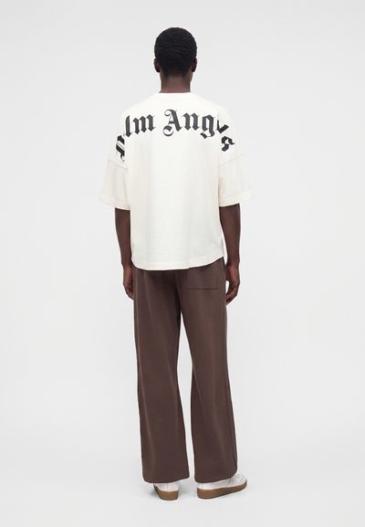 Άνδρας με πλάτη φοράει λευκό oversized μπλουζάκι με μεγάλο μαύρο κείμενο στο πάνω μέρος της πλάτης και καφέ φαρδιά παντελόνια με λευκά παπούτσια.