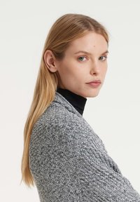 Blonde Frau mit hellblauen Augen in einem grauen Strickpullover und schwarzem Rollkragenpullover, die über ihre Schulter gegen einen schlichten hellen Hintergrund schaut.