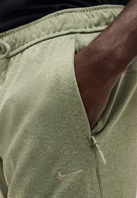 Δεξί χέρι που έχει τοποθετηθεί στην τσεπάκι με φερμουάρ ενός ανοιχτό πράσινου παντελονιού φόρμας Nike με ορατό κορδόνι μέσης.