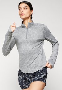 Graues, langärmliges Sportoberteil mit einem Viertelreißverschluss, strukturiertem Stoff und Daumenlöchern, kombiniert mit gemusterten schwarzen Shorts.