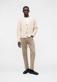 Μπεζ πλεκτό ζακέτα με κουμπιά, φορεμένο πάνω από ένα λευκό πουκάμισο. Συνδυασμένο με καφέ παντελόνι από κορντορό και μαύρα παπούτσια χωρίς κορδόνια.