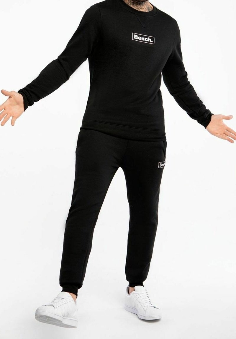 Homme portant un sweat-shirt Bench noir et un jogging avec des baskets blanches, debout les bras ouverts sur fond blanc.