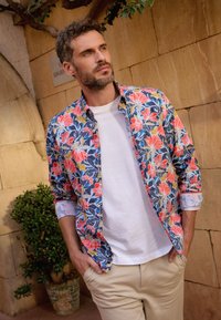 Bloemenoverhemd met blauwe, roze en groene patronen. Lichtgekleurde t-shirt met korte mouwen eronder. Beige broek. Gelaagde achtergrond.