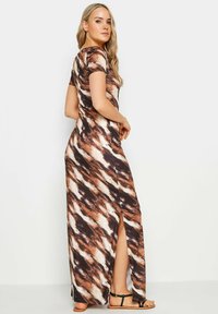 Robe maxi dans un motif tie-dye marron, blanc et noir ; manches courtes ; fente sur le côté ; tissu lisse ; associée à des sandales noires.