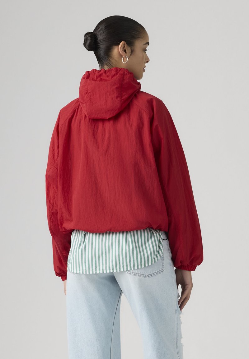 Frau mit Haaren zu einem Dutt zusammengebunden trägt eine rote Kapuzenjacke über einem grün gestreiften Hemd und hellblauen Jeans, steht mit dem Rücken zum Betrachter vor einem schlichten grauen Hintergrund.