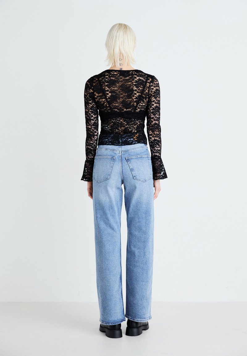 Haut en dentelle noire à manches longues, détail au dos visible. Jean large bleu clair avec poches classiques et finition délavée. Bottes noires chunky.