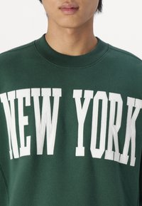 Green sweatshirt featuring large white letters spelling "NEW YORK" across the chest. Made of soft fabric with a round neckline.