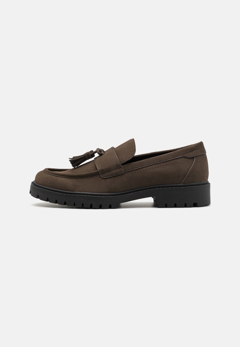 Mocassin en daim marron avec détail pompon, semelle épaisse en caoutchouc noir et petit talon, vu de profil sur un fond uni.