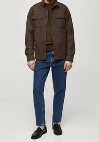 Chaqueta de lana marrón con dos bolsillos en el pecho, llevada sobre un suéter marrón, combinada con vaqueros azules y mocasines negros. Diseño sencillo y casual.