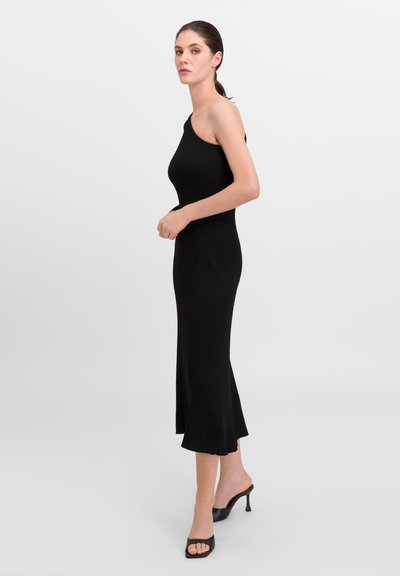 Robe noire à côtes avec un design à une épaule, silhouette ajustée et ourlet évasé. Portée avec des talons ouverts noirs.