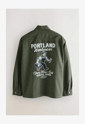 Olívazöld munkadzseki gombos gallérral, a hátán fehér grafika, amely egy embert, szerszámosládát és vastag betűs szöveget ábrázol.