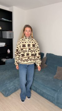 Pulover iz flisa z visokim ovratnikom, v kremni barvi z geometričnimi rjavimi vzorci. Oblečen z modrimi zvonastimi kavbojkami.