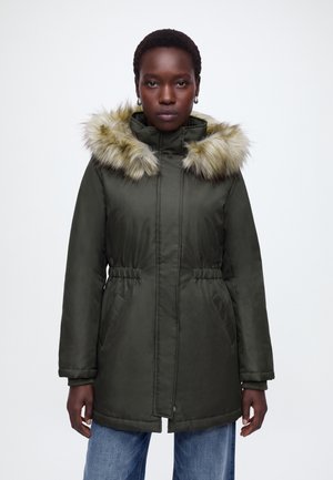Femme portant un manteau d'hiver vert foncé avec une capuche doublée de fourrure et un jean bleu, debout devant un fond clair uni.