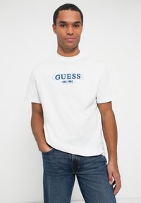 Hombre vistiendo una camiseta blanca de Guess con texto del logo azul y jeans azules, de pie con una mano en el bolsillo contra un fondo blanco liso.