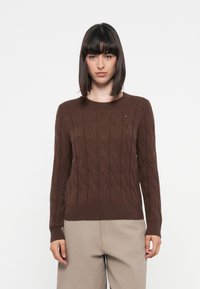 Brown knitted sweater featuring a textured cable pattern, round neck, and ribbed cuffs and hem. Subtle logo accent in the chest area.