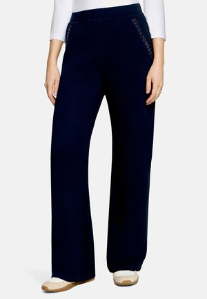 Femme portant un pantalon noir à jambes larges avec des poches latérales cloutées et des baskets blanches, montrée de la taille aux pieds sur un fond uni.