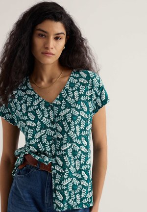 Young woman with long curly hair wearing a green short-sleeve blouse with white leaf print, tucked into blue jeans with a brown belt.