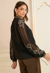 Melna blūze ar caurspīdīgām garām piedurknēm, kas rotāta ar ziedu izšuvumiem un pērļu akcentiem. Ribotie piedurkņu gali ar rakstainu detaļu ap apkakli.