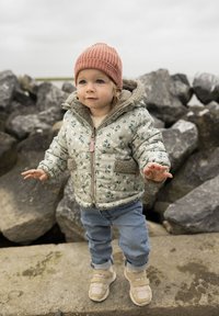 Vadderad ljusgrön jacka med blommönster, grå fleecefoder, tan accenter, ljusblå jeans, beige sneakers och en ribbad rosa beanie.