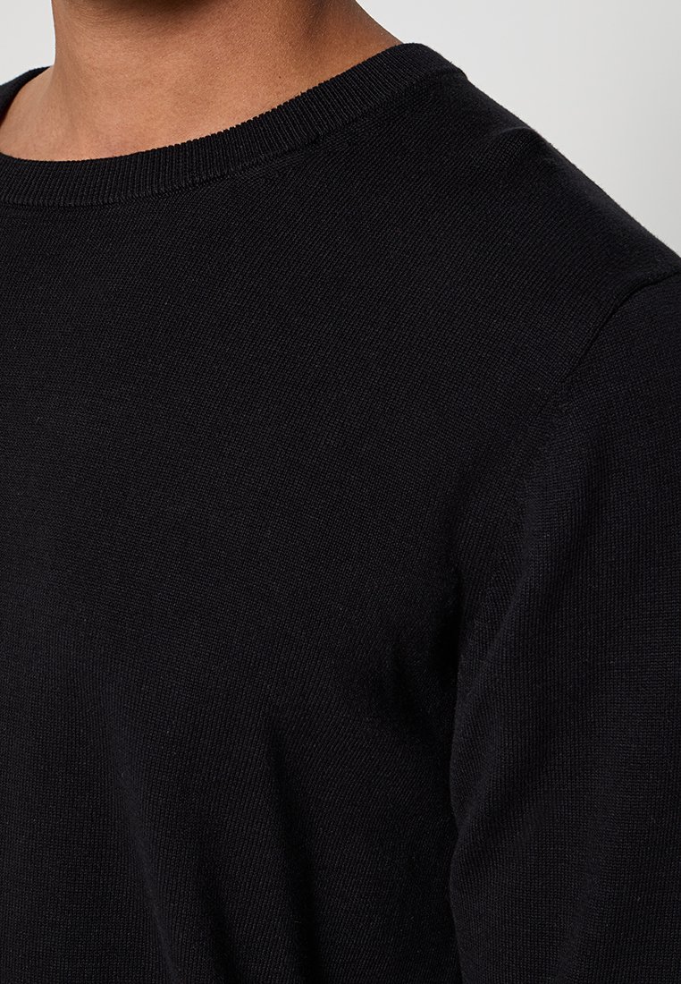 Primer plano de una persona que lleva un suéter de punto negro de manga larga con cuello redondo, mostrando la parte superior del pecho y el hombro.