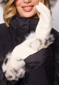 Białe, prążkowane rękawiczki dziane z akcentami z sztucznego futra w odcieniach kremu i brązu, noszone z czarną błyszczącą kurtką puchową.