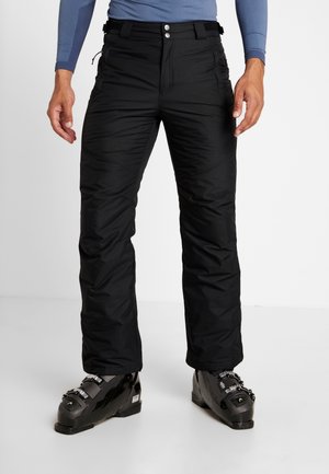 Mann trägt schwarze, isolierte Skihose und schwarze Skischuhe, steht vor einem schlichten weißen Hintergrund.