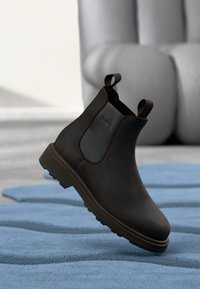 Braune Leder Chelsea-Boots mit elastischen Seitenpaneelen, Zuglaschen und strukturiertem Absatz, auf einem blauen Teppich präsentiert. Glatte Oberfläche und minimalistisches Design.