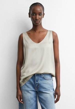 Donna con capelli intrecciati che indossa una canotta beige chiaro e jeans blu, in piedi davanti a uno sfondo bianco semplice.