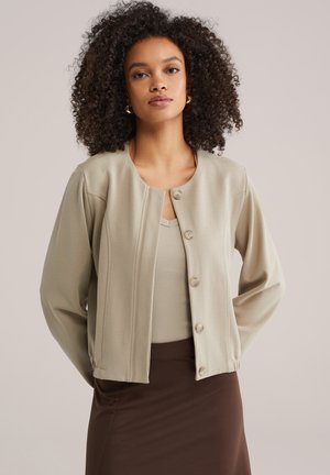 Hellbeige Jacke mit rundem Ausschnitt, langen Ärmeln und Knopfverschluss. Weiches Material mit glatter Textur, getragen über einem beigen Top und braunem Rock.