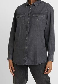 Camicia in denim grigio con maniche lunghe, tasche frontali, chiusura con bottoni e orlo leggermente curvo. Presenta cuciture a contrasto e bottoni in metallo.