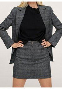 Blazer grigio a quadri con rever a punta, abbinato a un top nero aderente e a una gonna a tubino alta coordinata. Tessuto dalla texture liscia.