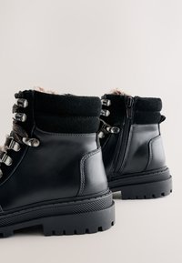 Black leather ankle boots with a rugged sole, suede accents, metal eyelets, and a zip closure at the back. Lined with soft brown material.