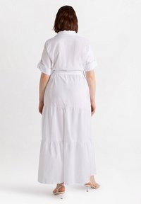 Robe maxi blanche avec un design à volants, manches courtes retroussées et taille ceinturée. Tissu lisse, détails minimaux et sans motifs visibles.