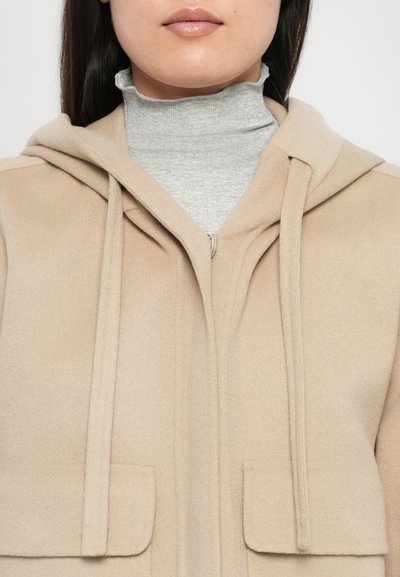 Veste à capuche beige en tissu doux avec deux poches avant, des détails de couture subtils et une fermeture éclair sur le devant. Pull à col roulé gris en dessous.