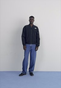 Giacca bomber nera con zip, dotata di logo, abbinata a jeans baggy azzurri e sneakers nere su un pavimento blu.