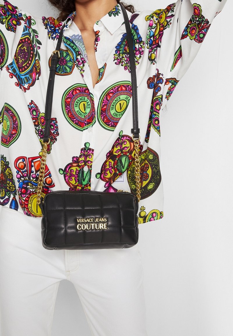 Czarna pikowana torba na ramię ze złotym okuciem, z łańcuszkowym paskiem i wytłoczonym napisem "VERSACE JEANS COUTURE" z przodu, trzymana na tle kolorowej koszuli.