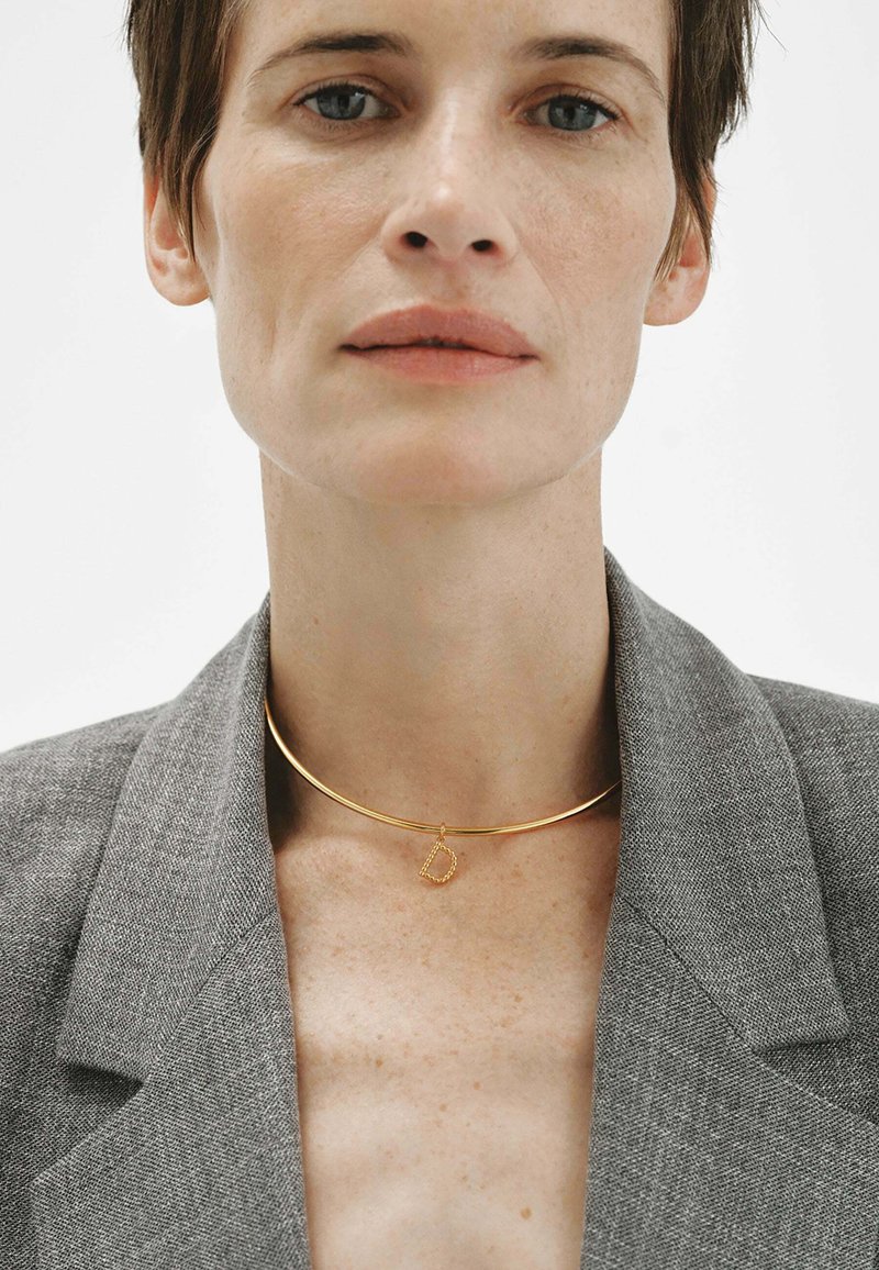 Persona con un blazer gris y un collar choker dorado con un colgante en forma de letra "D" sobre un fondo blanco.