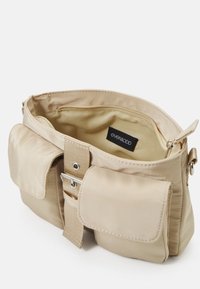 Beige tyg axelväska med två yttre fickor, metallbeslag och ett dragkedjeslutet huvudfack. Inuti finns en logomärke.