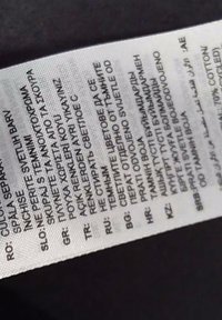 Etichetta di cura in tessuto bianco con più lingue, con testo nero che dettaglia le istruzioni di lavaggio e avvertenze sul colore. Superficie testurizzata, forma rettangolare.