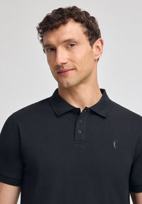 Svart polotröja i bomull, med klassisk krage, treknappslå och en liten broderad logotyp på bröstet. Slät textur.