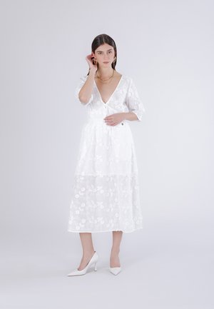 Nainen seisoo valkoisessa kukallisessa pitsissa tehdyssä midimekossa, jossa on 3/4-hihat, ja valkoisissa kapeakärkisissä korkeissa koroissa, yksiväristä vaaleaa taustaa vasten.