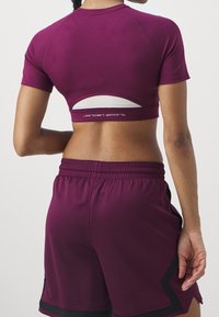 Žena v fialovém zkráceném sportovním topu s průstřihem a odpovídajících fialových sportovních šortkách s černými lemy, zachycena zezadu.