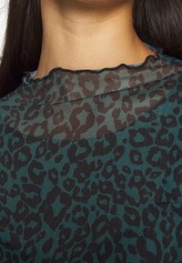 Transparent topp med ett mönster av mörkgrön och svart leopardprint, med en vågformad halsringning och textilstruktur.